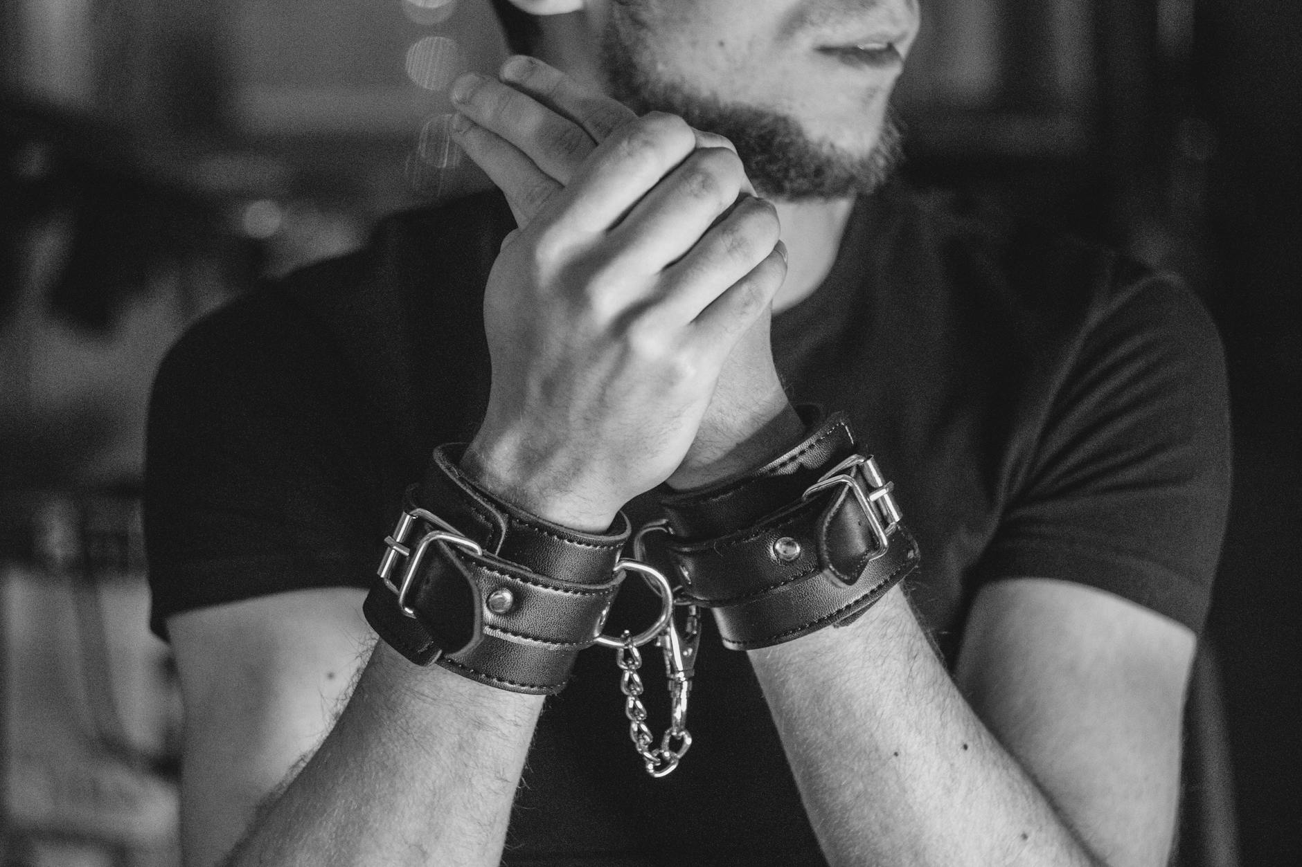 Close-up of a man's hands in leather cuffs, evoking themes of bondage and restraint.