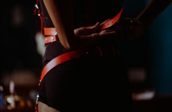 Back view of crop unrecognizable female in black corset and panties fastening leather harness in dressing room