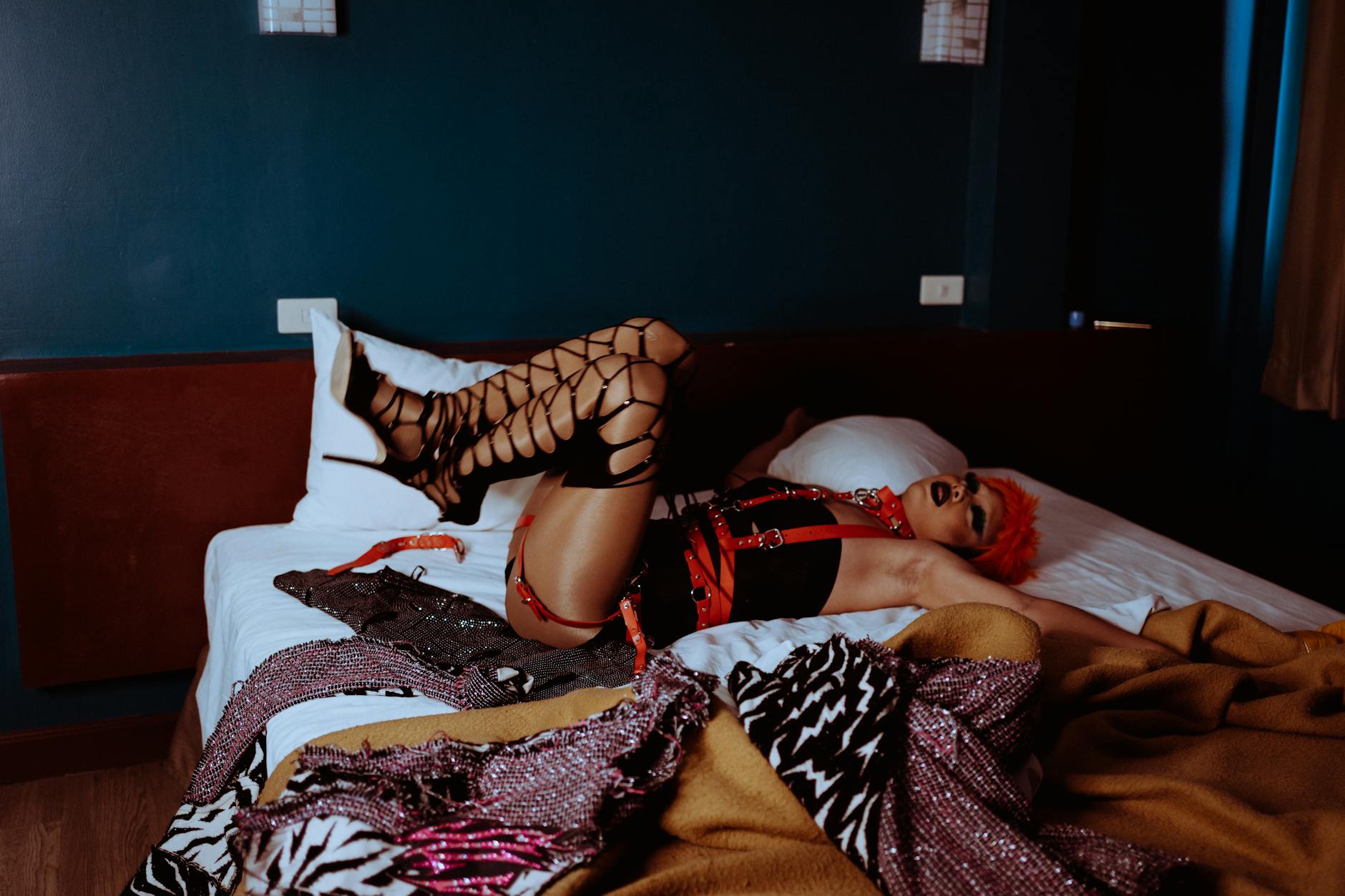 Full body slim male model with funky makeup and orange short hair in BDSM bodysuit and lace up shoes lying with legs raised on comfy bed and looking away