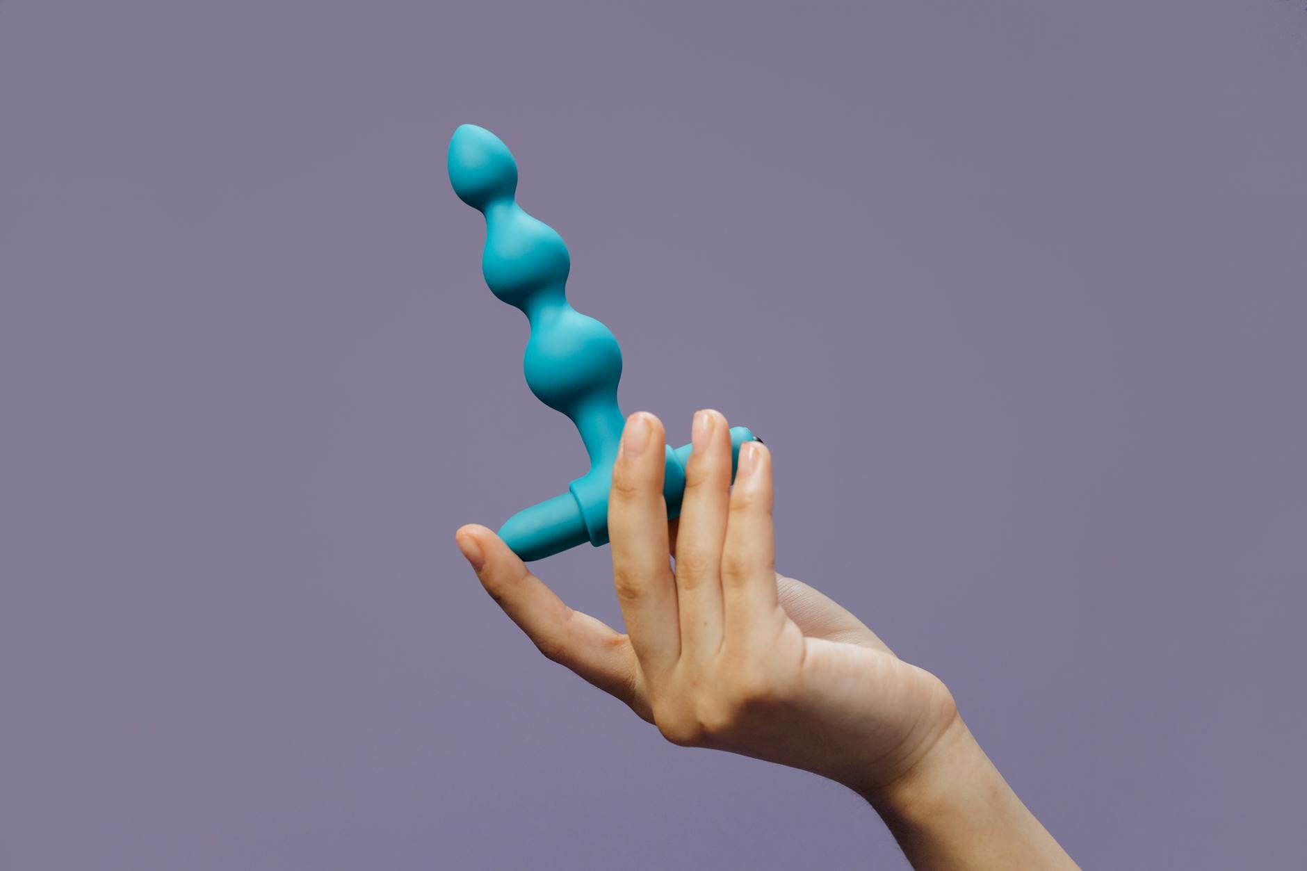 Close-up of a hand holding blue silicone anal beads against a purple background highlighting sensuality and self-love.