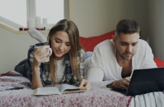 Cheerful couple have rest at home. Young man and woman lying in bed reading book and using laptop computer in the morning