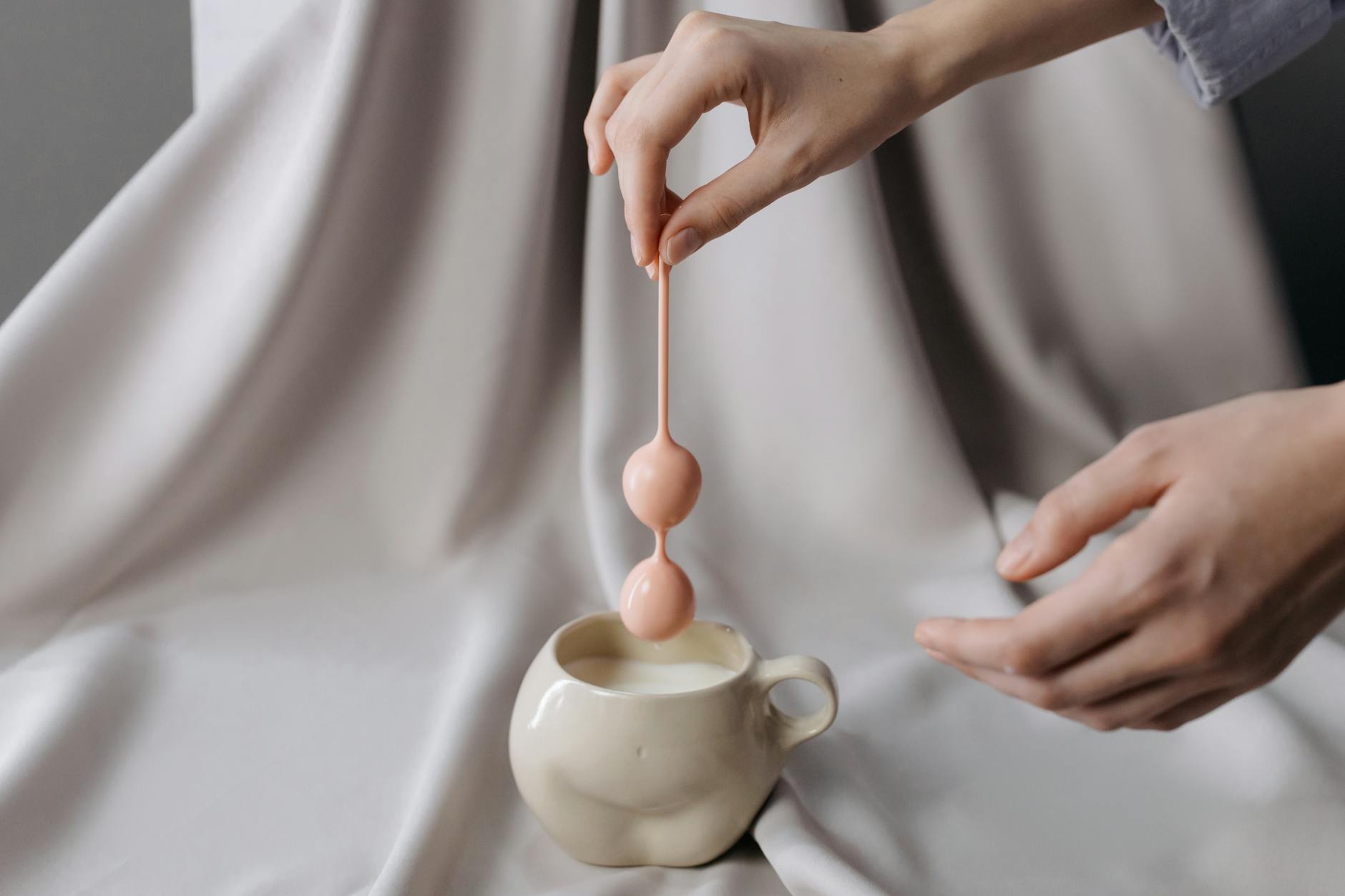 An artistic pastel scene featuring hands holding kegel balls over a mug of liquid