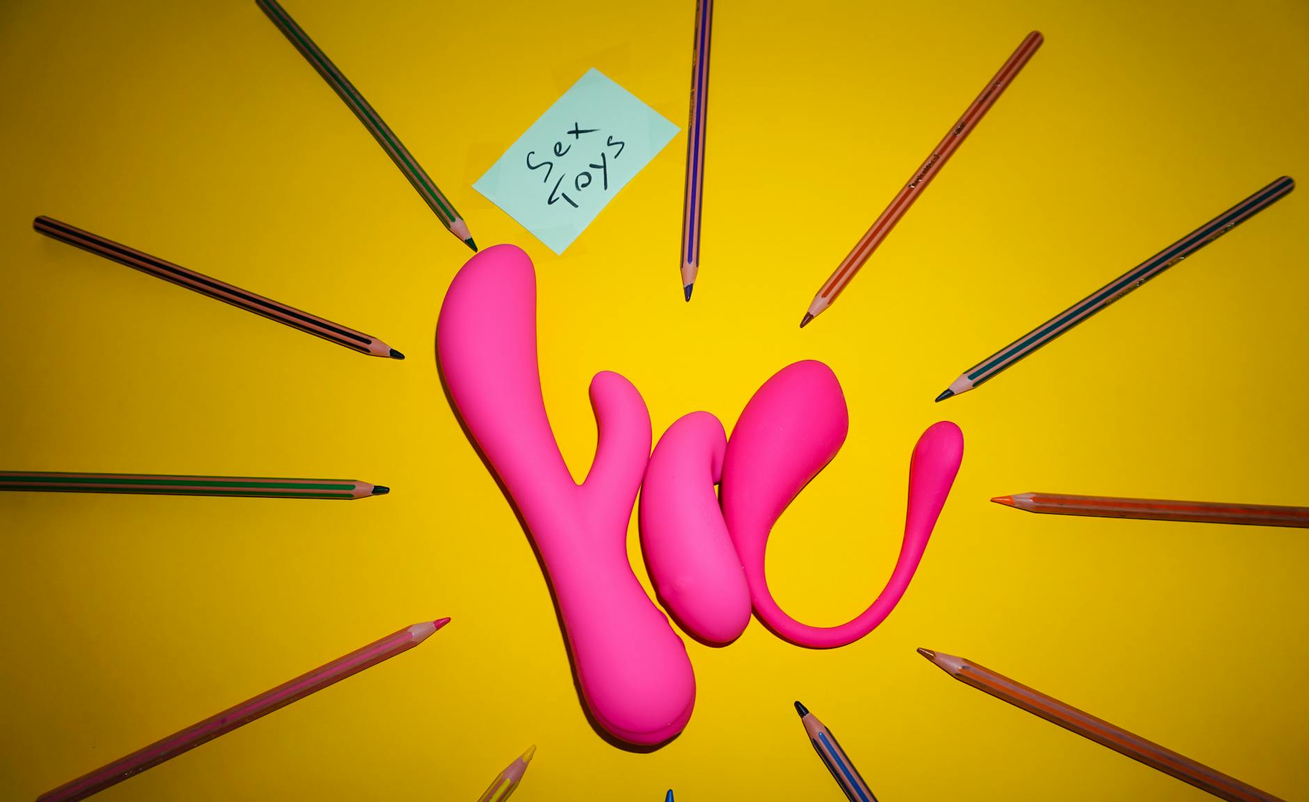 Pink sex toys surrounded by colored pencils on a yellow background