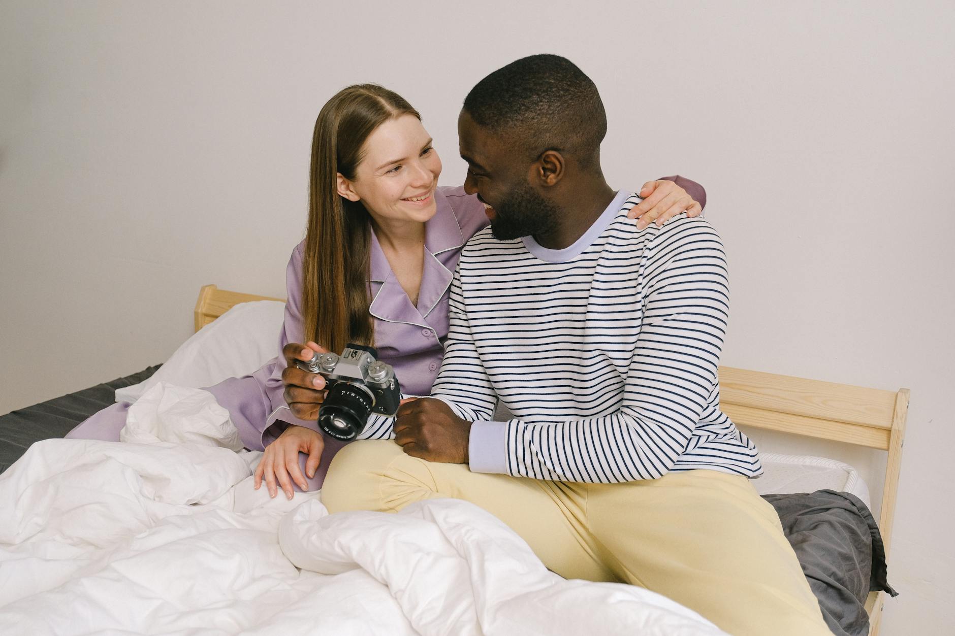 Cheerful multiracial couple looking at each other while sitting on comfortable bed with photo camera in light bedroom at home