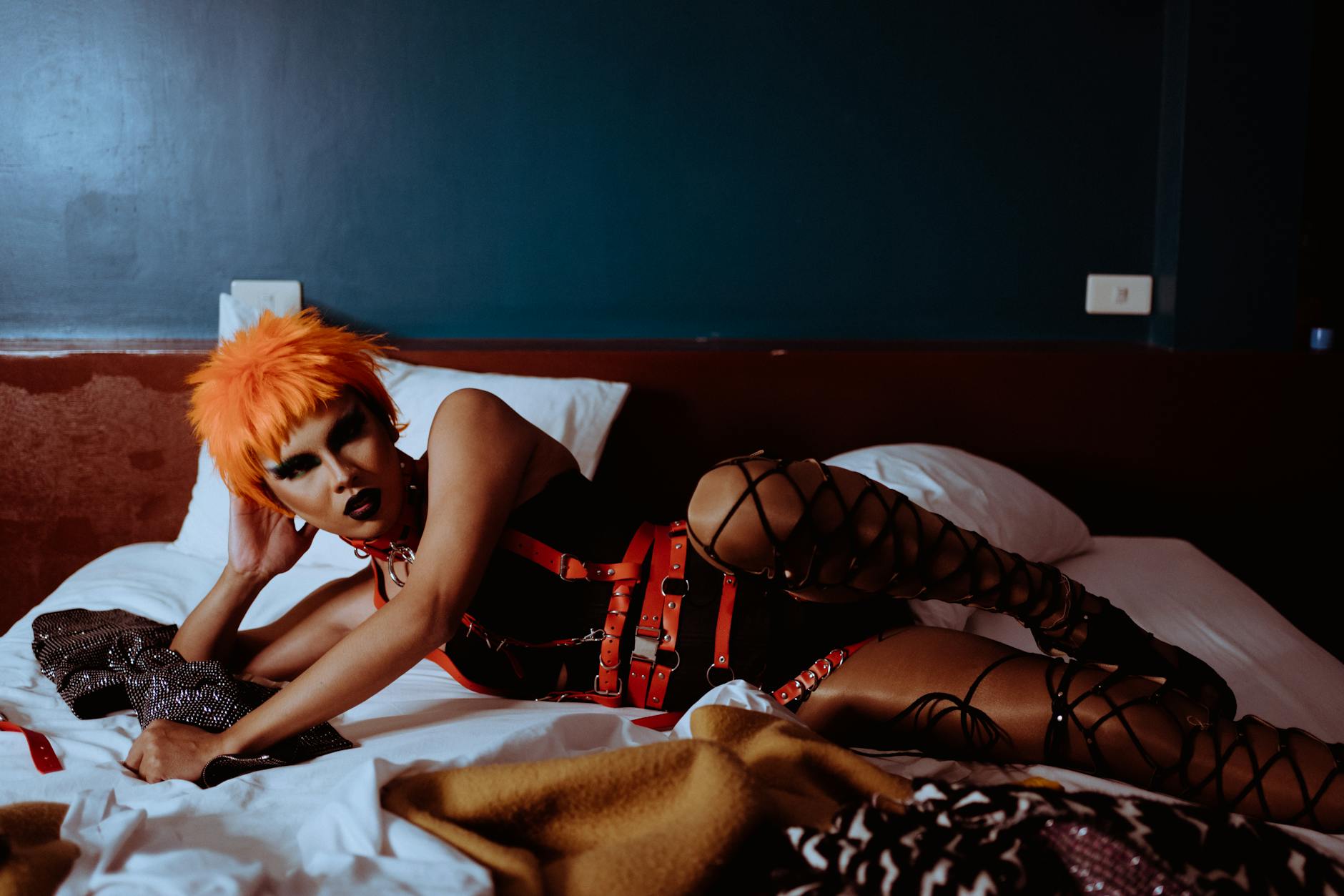 From above of eccentric young ethnic prostitute in with dark makeup and wig in bodysuit and BDSM harness lying on bed leaning on hand and looking at camera